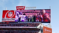A large screen at Levi's Stadium displays a football game with players from two teams, likely in a pre-game or mid-game moment. The screen prominently features the NFL logo and the word 'DIVISION'. The screen is above a crowded stadium with the audience visible below. Advertisements for sponsors like Dignity Health and Mountain Mike's Pizza are also noticeable in the scene.