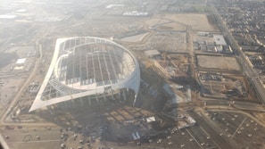 An aerial view of a large, partially constructed stadium with a distinctive oval shape. The area surrounding the structure is filled with construction materials and equipment, as well as parking lots. Several cranes are visible, suggesting ongoing work. The surrounding landscape is urban, with visible roads and buildings.