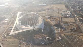 An aerial view of a large, partially constructed stadium with a distinctive oval shape. The area surrounding the structure is filled with construction materials and equipment, as well as parking lots. Several cranes are visible, suggesting ongoing work. The surrounding landscape is urban, with visible roads and buildings.