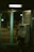 A dimly lit phone booth contains a vintage-style payphone attached to the wall. The booth has teal-colored metallic frames and glass windows with reflections. The outside environment is dark with blurred street lights in the background, suggesting it is nighttime.