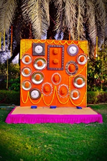 An elaborately decorated backdrop featuring multiple clocks and ornate frames arranged symmetrically on an orange panel, adorned with yellow marigold flowers and garlands. The setup is placed on a pink-draped platform, with lush foliage and palm fronds visible in the background.