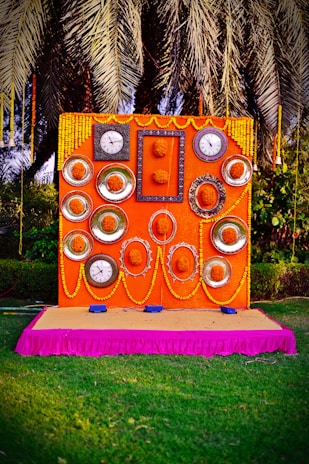 An elaborately decorated backdrop featuring multiple clocks and ornate frames arranged symmetrically on an orange panel, adorned with yellow marigold flowers and garlands. The setup is placed on a pink-draped platform, with lush foliage and palm fronds visible in the background.