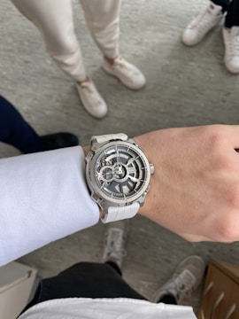 A close-up view of a wristwatch with a complex, skeleton-style face, worn on a person's wrist. The watch has a white strap and is positioned against a background where multiple people are visible wearing casual footwear on a grey floor.