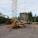 A construction site features a mobile crane with a tall metal lattice structure, lifting large concrete slabs. The area is surrounded by trees and residential buildings, with scattered equipment and materials on the gravel ground.