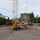 A construction site features a mobile crane with a tall metal lattice structure, lifting large concrete slabs. The area is surrounded by trees and residential buildings, with scattered equipment and materials on the gravel ground.