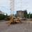 A construction site features a mobile crane with a tall metal lattice structure, lifting large concrete slabs. The area is surrounded by trees and residential buildings, with scattered equipment and materials on the gravel ground.