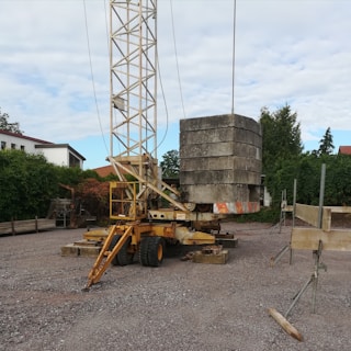 A construction site features a mobile crane with a tall metal lattice structure, lifting large concrete slabs. The area is surrounded by trees and residential buildings, with scattered equipment and materials on the gravel ground.
