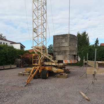 A construction site features a mobile crane with a tall metal lattice structure, lifting large concrete slabs. The area is surrounded by trees and residential buildings, with scattered equipment and materials on the gravel ground.