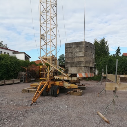 A construction site features a mobile crane with a tall metal lattice structure, lifting large concrete slabs. The area is surrounded by trees and residential buildings, with scattered equipment and materials on the gravel ground.