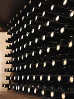 A neatly organized wine cellar with rows of wine bottles stored horizontally on wooden shelves. Each bottle is uniformly placed with corks facing outward, creating a visually appealing pattern. The cellar's ambient lighting casts a warm glow over the scene.