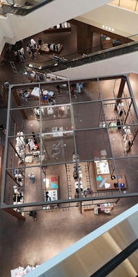 A multi-level indoor shopping mall with an open design and escalators visible. The image captures the bird's-eye view of various sections with organized displays of clothing and accessories. Shoppers are browsing through the store, creating a bustling yet organized atmosphere.