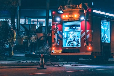 A fire truck is parked on a dimly lit street at night with its compartments open and equipment visible. Several firefighters in reflective gear are standing nearby, including one seen walking towards the vehicle. The scene is illuminated by the lights from the fire truck and streetlights, creating a contrast between the red and blue lights.