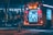 A fire truck is parked on a dimly lit street at night with its compartments open and equipment visible. Several firefighters in reflective gear are standing nearby, including one seen walking towards the vehicle. The scene is illuminated by the lights from the fire truck and streetlights, creating a contrast between the red and blue lights.