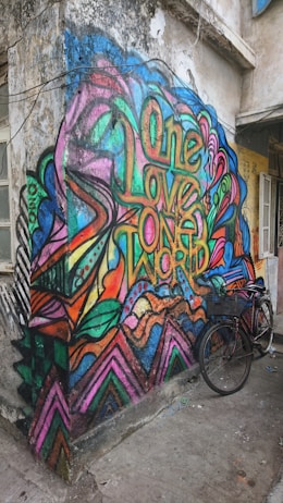 A vibrant and colorful graffiti mural on a worn-down urban wall, featuring abstract patterns and the words 'One Love One World' in bold, stylized letters. A bicycle is parked next to the mural, adding an element of everyday life to the lively street art scene.