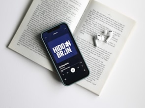 A smartphone displaying a podcast screen titled 'Hidden Brain' lies on an open book. Nearby, a pair of white wireless earbuds rests on the pages. The scene suggests a moment of leisure or learning, combining technology with traditional reading.