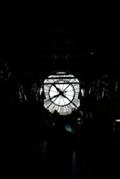 A large, backlit clock with Roman numerals is visible, silhouetted against a bright background. Several people stand in shadow in front of the clock, creating a mysterious and dramatic scene.