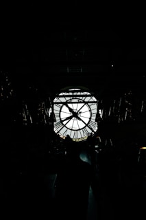 A large, backlit clock with Roman numerals is visible, silhouetted against a bright background. Several people stand in shadow in front of the clock, creating a mysterious and dramatic scene.
