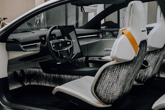 A modern car interior featuring a sleek steering wheel with a digital display behind it. The dashboard has minimalist lines with integrated technology, including a touch screen. The seats are white with black mesh backing and accented by an orange safety belt.