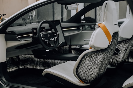 A modern car interior featuring a sleek steering wheel with a digital display behind it. The dashboard has minimalist lines with integrated technology, including a touch screen. The seats are white with black mesh backing and accented by an orange safety belt.