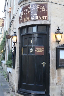 A charming entrance to a hotel and restaurant with a dark wooden door adorned with a decorative metal face. The entrance has a vintage sign above it reading 'Candlesticks Hotel and Restaurant.' Two traditional lanterns flank the door, providing a warm, inviting glow. Potted greenery lines the stone building, adding a touch of nature to the scene.