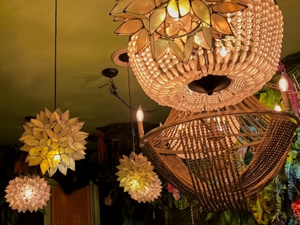 Elegant chandeliers with intricate designs hang from the ceiling, composed of delicate beads and petals that create a warm, inviting glow. The surroundings feature lush greenery and floral decorations that add a natural ambiance to the scene.