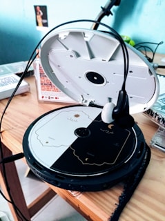 A portable CD player is open on a wooden surface, revealing a disc with a black and white design featuring text and small icons. Headphones are placed over the player. In the background, there are various items including a box with text and a small visible photograph on the wall.
