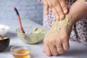 A person applying a green paste, possibly a homemade mask or scrub, on their hand. In the background, an open avocado, a bowl containing more of the green mixture, a spoon with an orange handle, and a small bowl of honey are visible.
