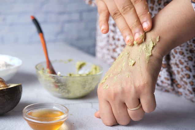 A person applying a green paste, possibly a homemade mask or scrub, on their hand. In the background, an open avocado, a bowl containing more of the green mixture, a spoon with an orange handle, and a small bowl of honey are visible.