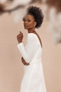 A person with natural curly hair and elegant jewelry stands with a confident pose. The individual wears a white dress with a deep V-back, set against a soft, neutral background. Their expression appears calm and poised.