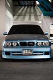 A blue car with a BMW M3 emblem is parked indoors under industrial lighting. Its headlights and grille are distinct, and there are colorful pipes overhead.