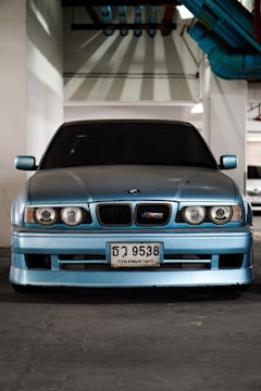 A blue car with a BMW M3 emblem is parked indoors under industrial lighting. Its headlights and grille are distinct, and there are colorful pipes overhead.