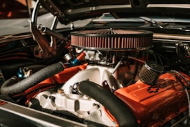 A detailed view of a car engine featuring a prominently visible air filter surrounded by various hoses and metal components. The engine has a bold Chevrolet logo and vibrant orange covers.