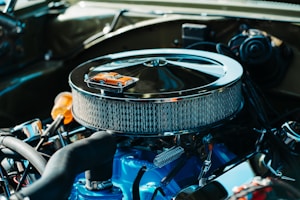 A detailed view of a car engine with a chrome air filter housing prominently displayed. On top of the air filter sits a miniature orange toy car, adding a whimsical element to the industrial setup. The engine components are clean and well-maintained, featuring a combination of mechanical parts in various materials.