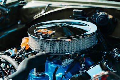 A detailed view of a car engine with a chrome air filter housing prominently displayed. On top of the air filter sits a miniature orange toy car, adding a whimsical element to the industrial setup. The engine components are clean and well-maintained, featuring a combination of mechanical parts in various materials.