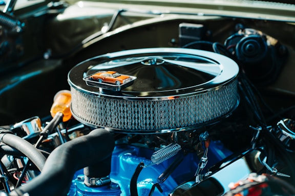 A detailed view of a car engine with a chrome air filter housing prominently displayed. On top of the air filter sits a miniature orange toy car, adding a whimsical element to the industrial setup. The engine components are clean and well-maintained, featuring a combination of mechanical parts in various materials.