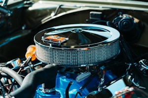 A detailed view of a car engine with a chrome air filter housing prominently displayed. On top of the air filter sits a miniature orange toy car, adding a whimsical element to the industrial setup. The engine components are clean and well-maintained, featuring a combination of mechanical parts in various materials.