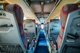A view down the aisle of a bus with colorful patterned seat covers and overhead storage compartments. The interior is well-lit, and the aisle is carpeted in blue. Seats have back pockets and adjustable armrests.