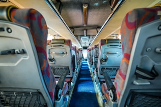 A view down the aisle of a bus with colorful patterned seat covers and overhead storage compartments. The interior is well-lit, and the aisle is carpeted in blue. Seats have back pockets and adjustable armrests.