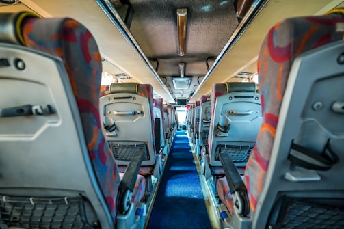A view down the aisle of a bus with colorful patterned seat covers and overhead storage compartments. The interior is well-lit, and the aisle is carpeted in blue. Seats have back pockets and adjustable armrests.