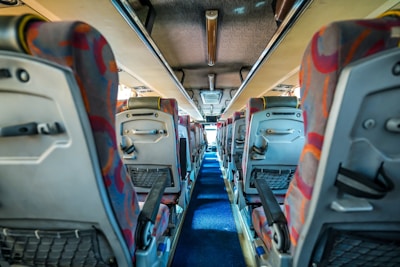 A view down the aisle of a bus with colorful patterned seat covers and overhead storage compartments. The interior is well-lit, and the aisle is carpeted in blue. Seats have back pockets and adjustable armrests.