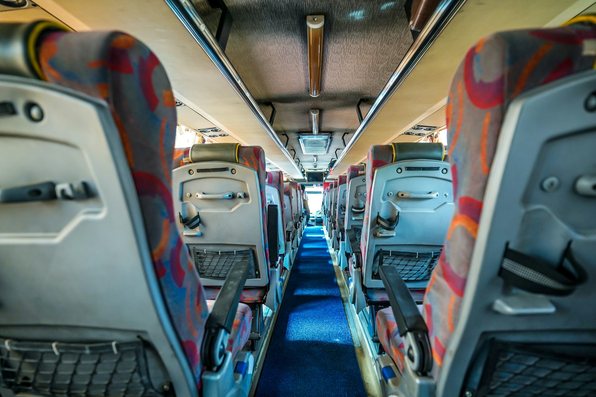 A view down the aisle of a bus with colorful patterned seat covers and overhead storage compartments. The interior is well-lit, and the aisle is carpeted in blue. Seats have back pockets and adjustable armrests.