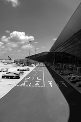 A black and white scene captures an airport environment with an airplane parked on the tarmac. Several service vehicles are aligned near the terminal, and the area is illuminated by natural daylight, casting shadows on the ground. In the background, large clouds are scattered across the sky.