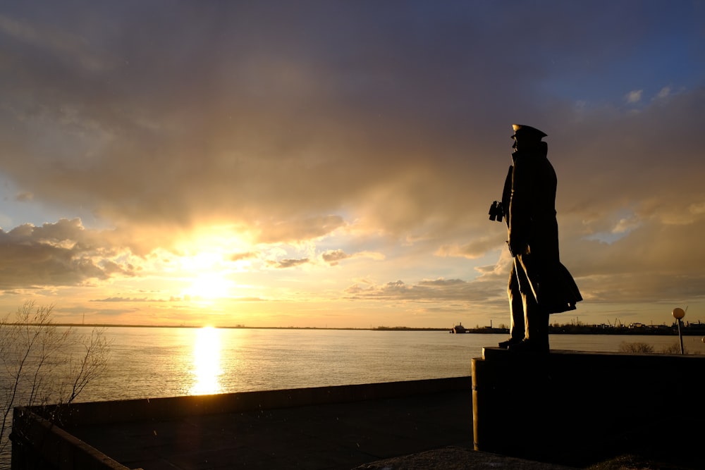 silhouette of woman standing on rock near body of water during sunset