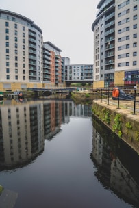Several modern high-rise residential buildings surround a small canal. The buildings feature large windows and balconies. The water reflects the structures, creating a mirrored image. A small walkway and bridge run between the edges of the canal, with some greenery and orange safety equipment visible along the railings.