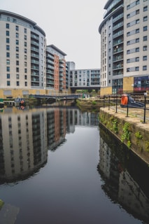 Several modern high-rise residential buildings surround a small canal. The buildings feature large windows and balconies. The water reflects the structures, creating a mirrored image. A small walkway and bridge run between the edges of the canal, with some greenery and orange safety equipment visible along the railings.