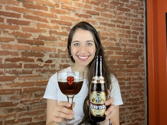 A person is holding a glass of dark beer in one hand and a bottle of Kvak beer in the other. They are smiling and standing against a backdrop of a textured red brick wall. The glass features the logo 'Old Monk Beer House'. The person is wearing a white shirt and has long dark hair.