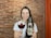 A person is holding a glass of dark beer in one hand and a bottle of Kvak beer in the other. They are smiling and standing against a backdrop of a textured red brick wall. The glass features the logo 'Old Monk Beer House'. The person is wearing a white shirt and has long dark hair.