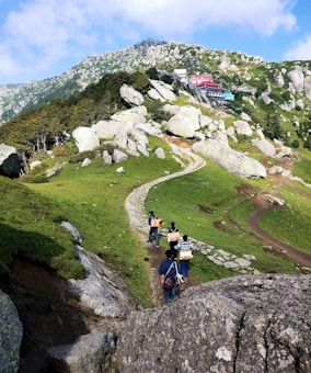 A scenic mountain pathway lined with rocks and greenery leads upward through a landscape with scattered trees and a hillside building in the distance. Several people walk along the path, carrying musical instruments and gear on their backs.