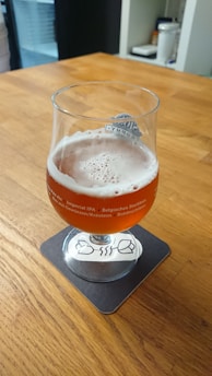 A half-full beer glass with an amber-colored beverage and a frothy head, placed on a coaster on a wooden table. The glass features text indicating various beer styles. In the background, there is a shelf with some items.