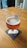 A half-full beer glass with an amber-colored beverage and a frothy head, placed on a coaster on a wooden table. The glass features text indicating various beer styles. In the background, there is a shelf with some items.
